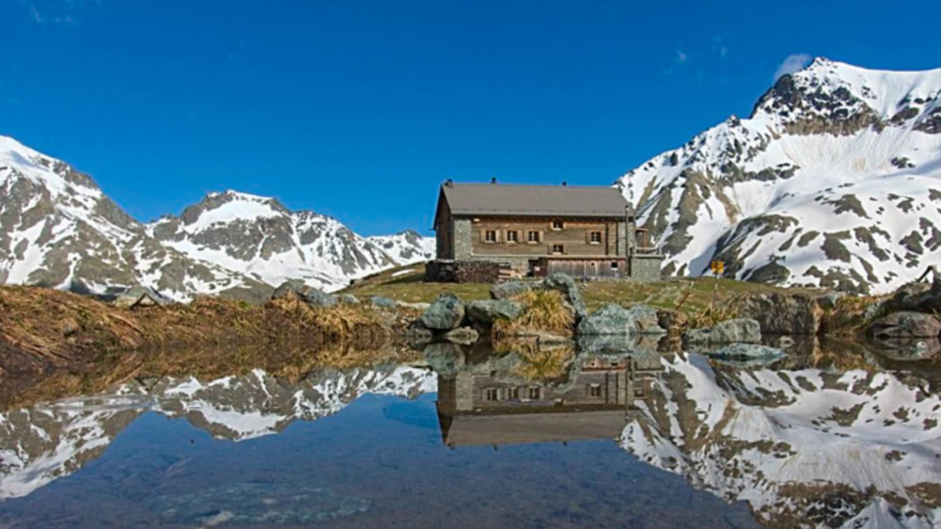Die Chamanna Jenatsch spiegelt sich perfekt in einem stillen Bergsee vor schneebedeckten Gipfeln wider.