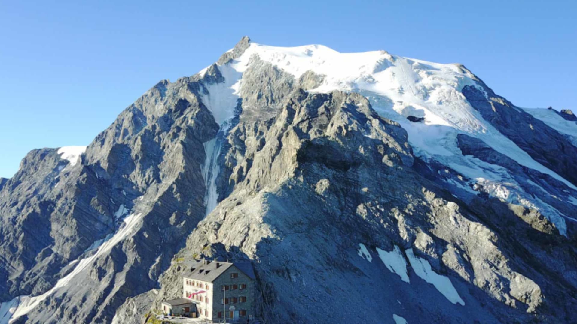 Luftaufnahme der Julius Payer Hütte auf einem schmalen Felsgrat mit verschneitem Gipfel und Gletscherzungen im Hintergru
