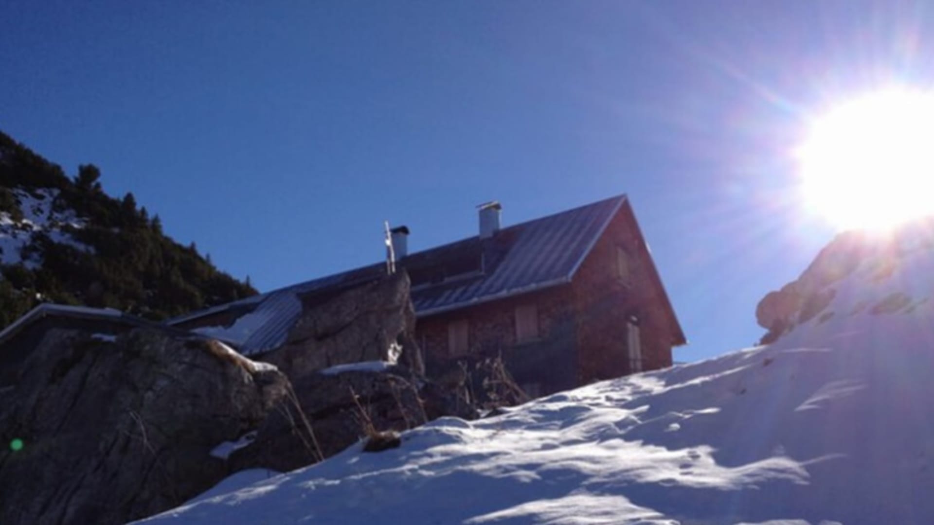 Die Freiburger Hütte aus Holz steht verschneit zwischen Felsen bei starkem Sonnenlicht im Winter.