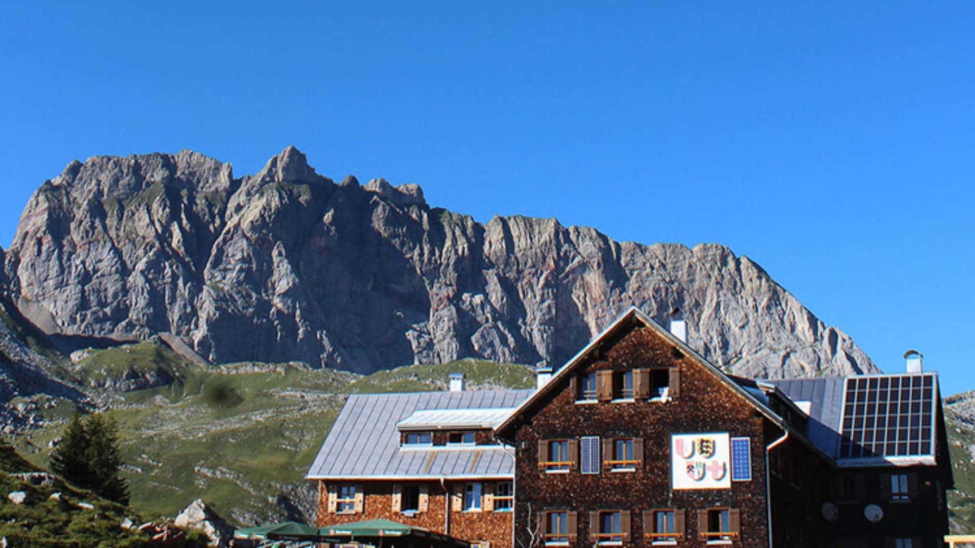 Die Freiburger Hütte mit Holzschindelfassade steht vor einer schroffen Felswand aus grauem Kalkstein.