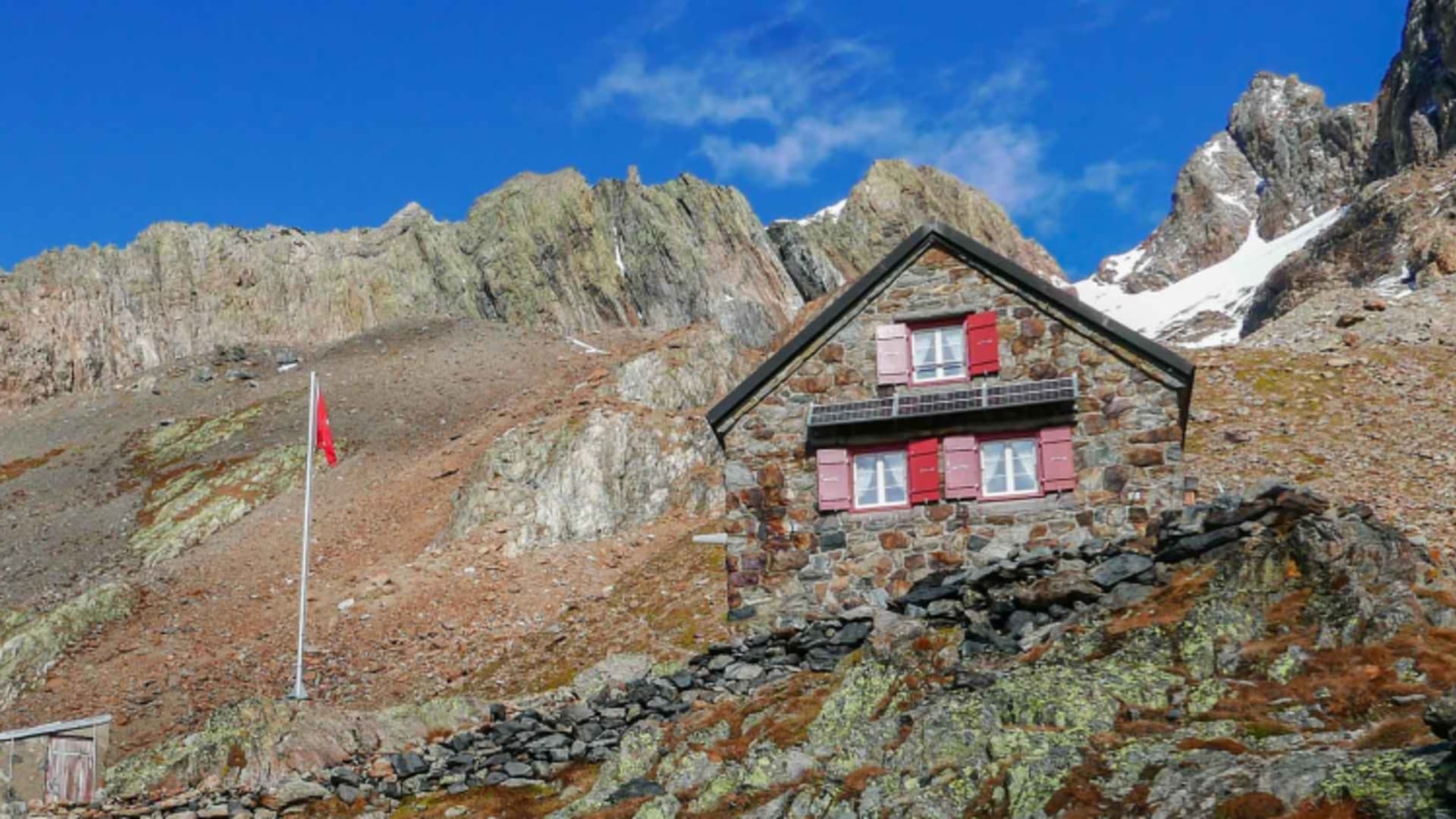 Die Trifthütte aus Naturstein mit rotem Holzdach steht auf felsigem Hang in hochalpiner Berglandschaft.
