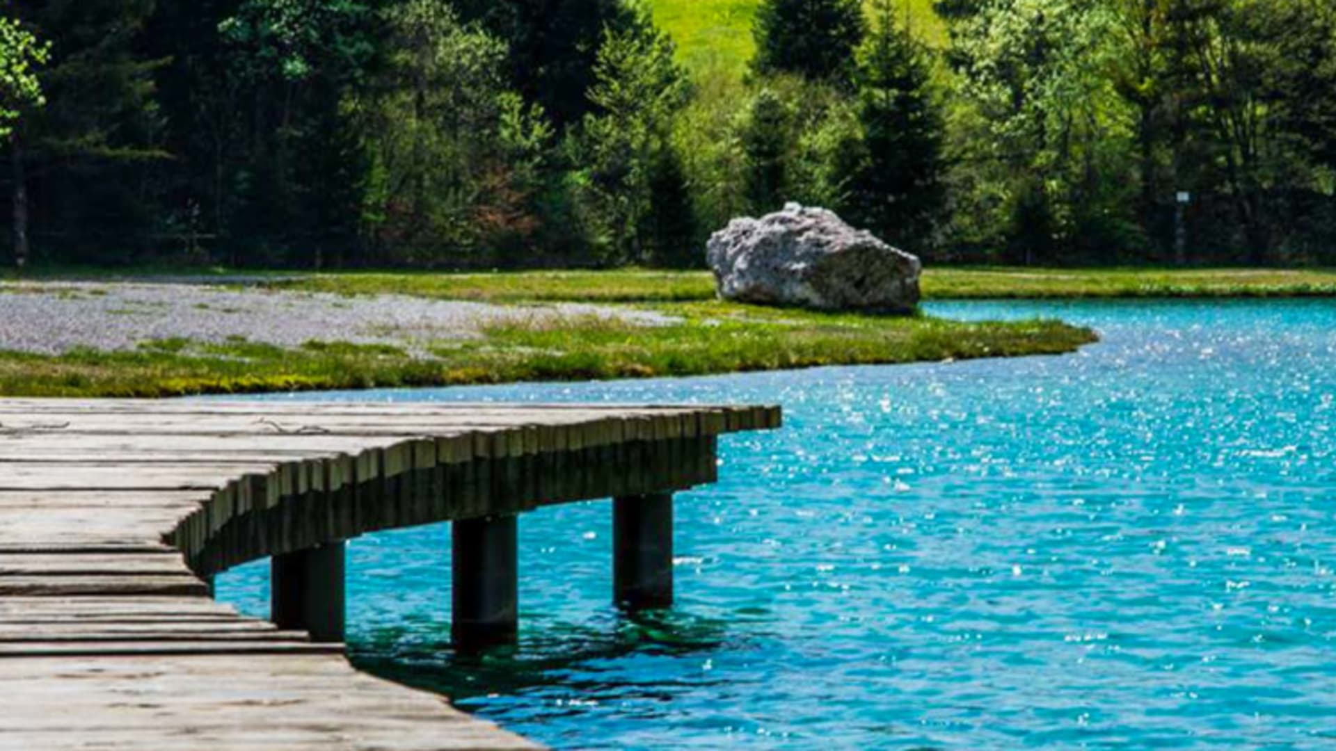 Holzsteg führt über kristallklares Wasser des Sees beim Garni Lago Nembia mit bewaldeten Ufern.