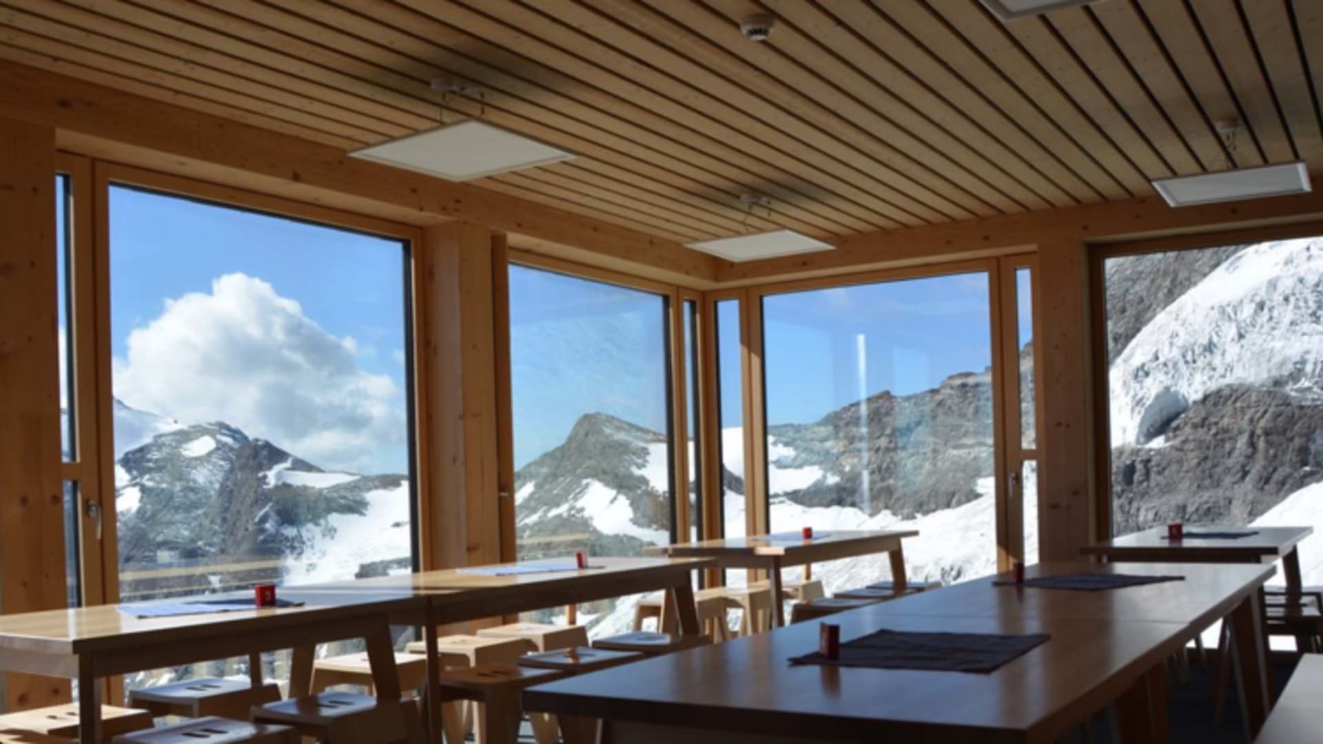 Heller Gastraum der Hörnlihütte mit bodentiefen Fenstern und Blick auf verschneite Gipfel der Bergkette.
