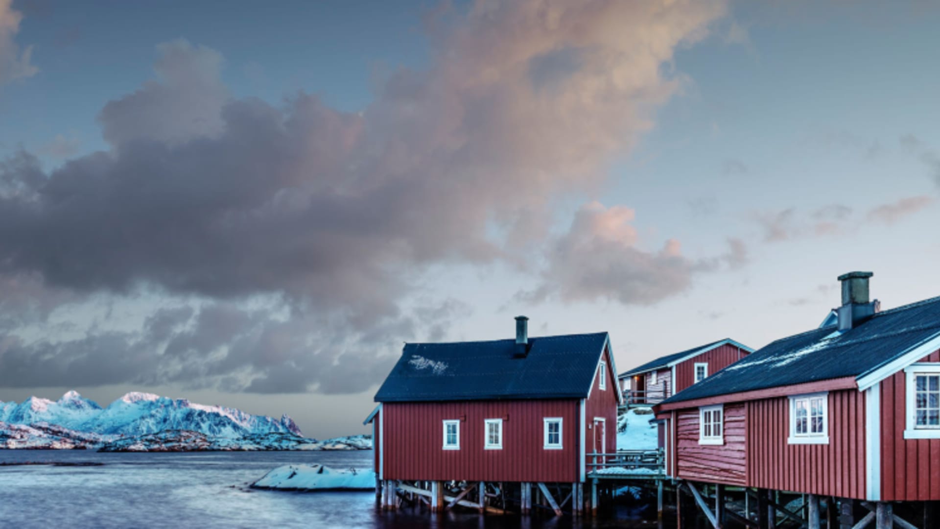Winterpanorama der Svinoya Rorbuer - rote Holzhütten auf Stelzen über dem Fjord mit verschneiten Bergen im Hintergrund.