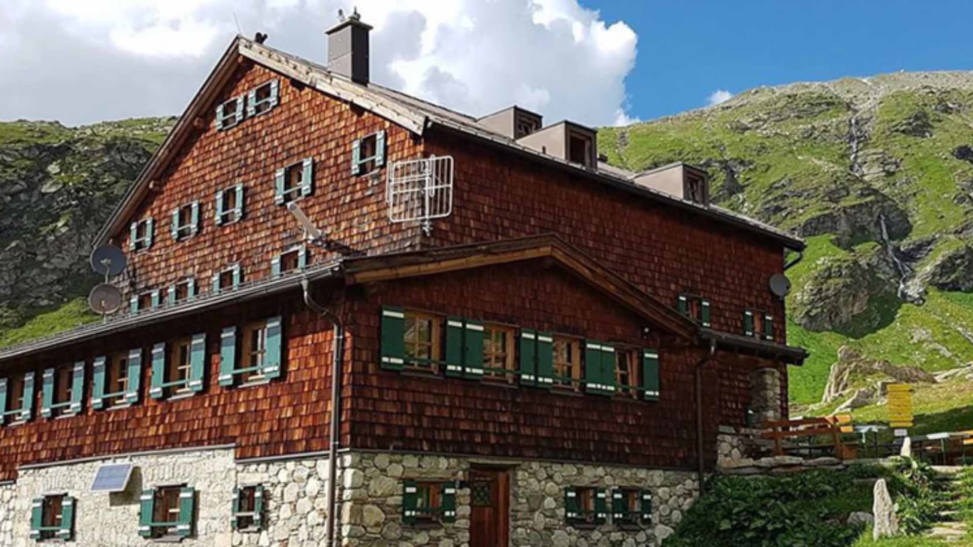 Haupthaus der Warnsdorfer Hütte mit Holzschindelfassade und Natursteinmauerwerk in grüner Almlandschaft.