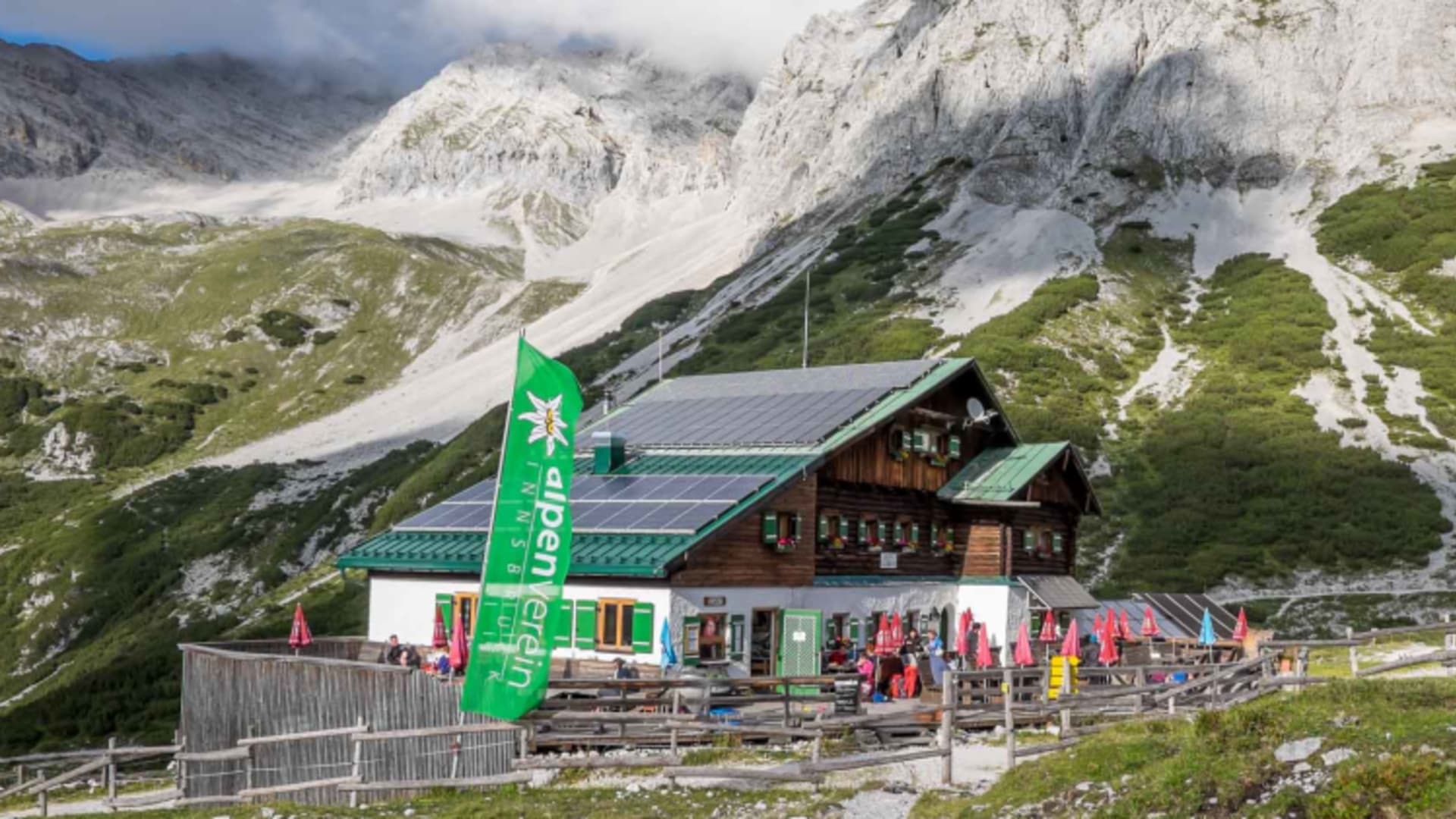 Bergsteiger auf der Terrasse der Pfeishütte unter der grünen Alpenvereins-Fahne mit Edelweiß.