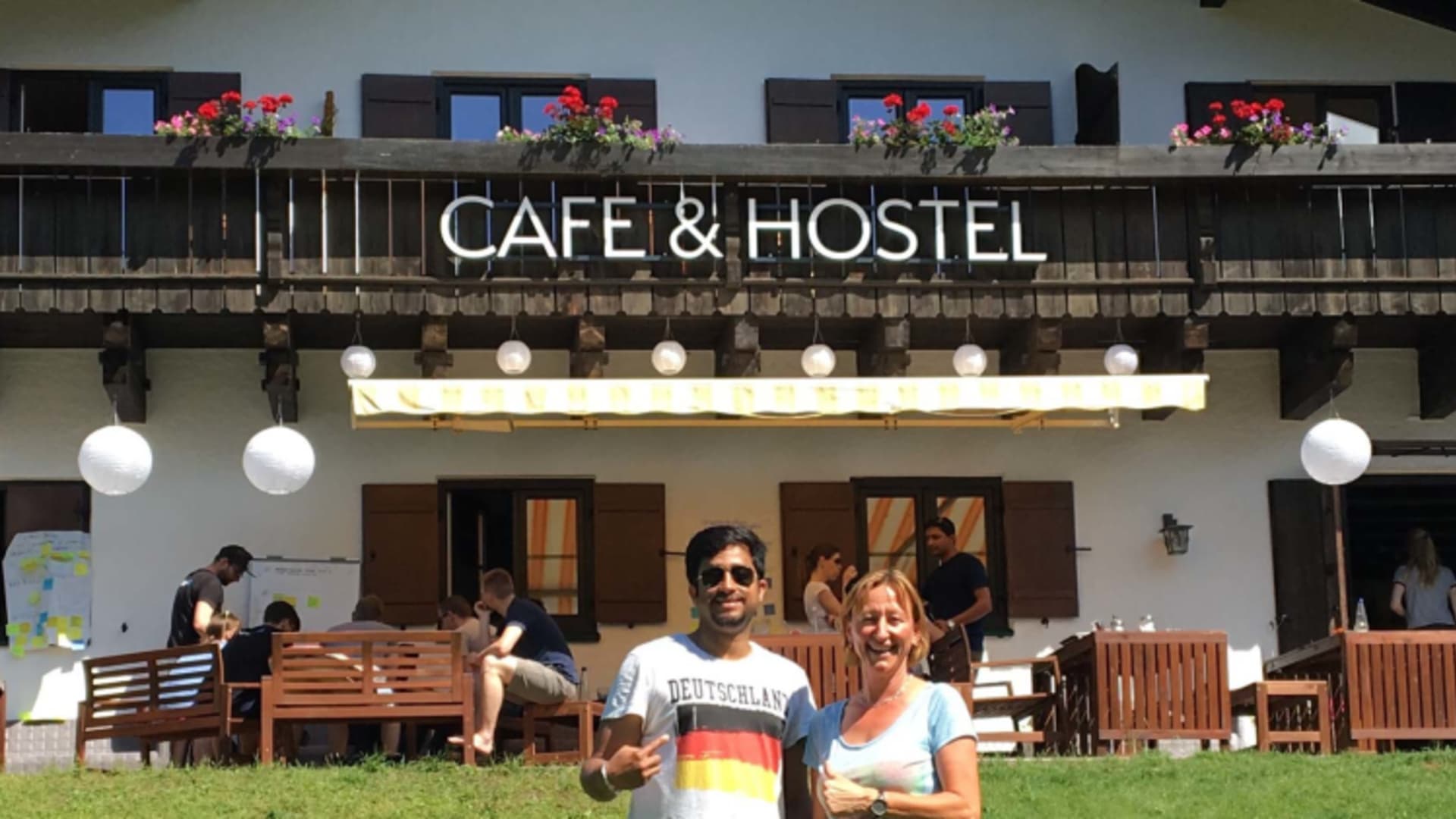 Pächter des Berg Blick Hostel posieren vor dem Café auf der Terrasse mit Holzmöbeln und Blumenschmuck.