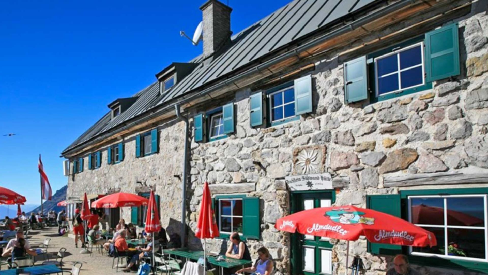 Wanderer pausieren auf der Sonnenterrasse der Dachstein Südwandhütte bei klarem Himmel im Hochgebirge.