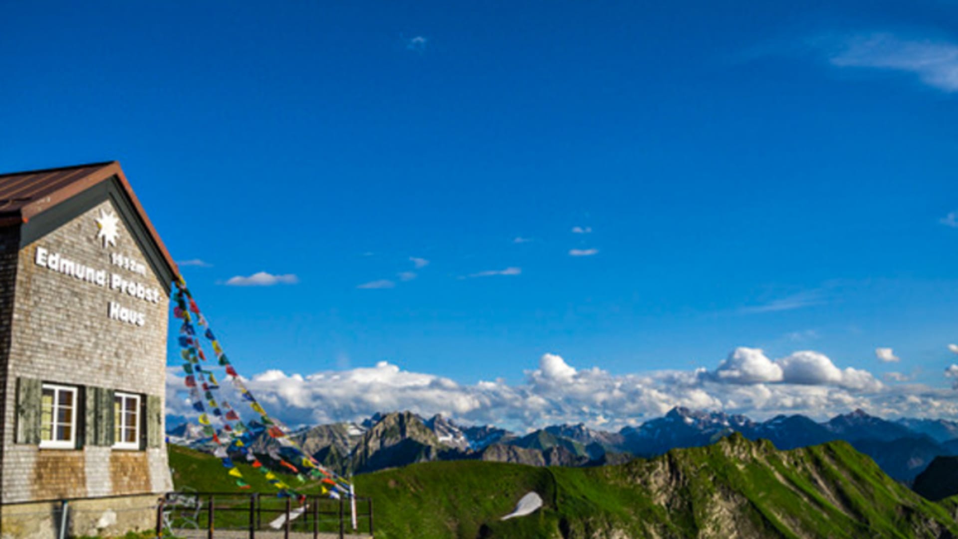 Außenansicht Edmund Probst Haus aus hellem Naturstein auf Bergkamm mit bunten Gebetsfahnen und Alpenpanorama im Hintergrund.