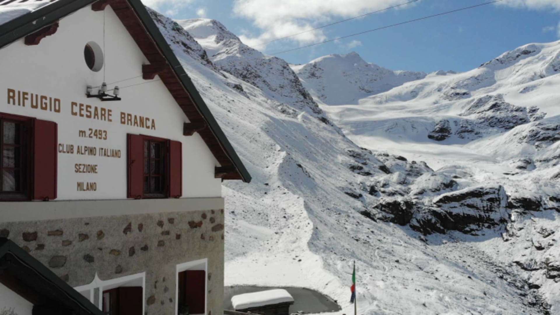 Außenansicht des Rifugio Branca mit Holzterrasse und italienischer Flagge vor verschneiter Bergkulisse.