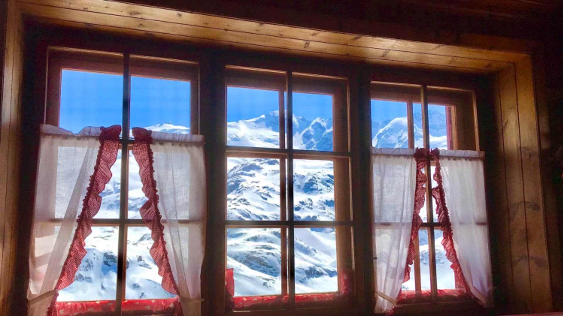 Durch die große Fensterfront der Gaststube im Rifugio Branca blickt man auf verschneite Bergkuppen unter blauem Himmel.