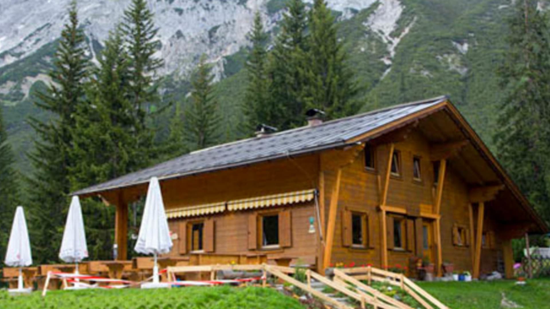 Die Neue Alplhütte steht auf grüner Bergwiese umgeben von dichtem Fichtenwald in Tirol.
