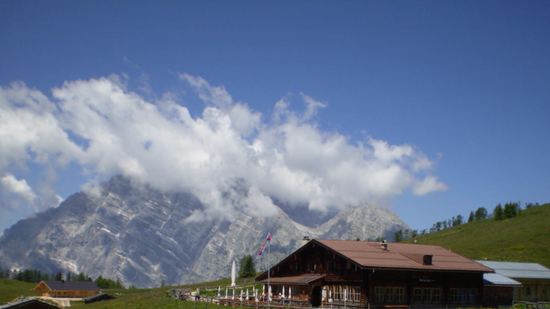 Die Gotzenalm als traditionelle Holzhütte mit Terrasse vor dem Bergpanorama der Umgebung.
