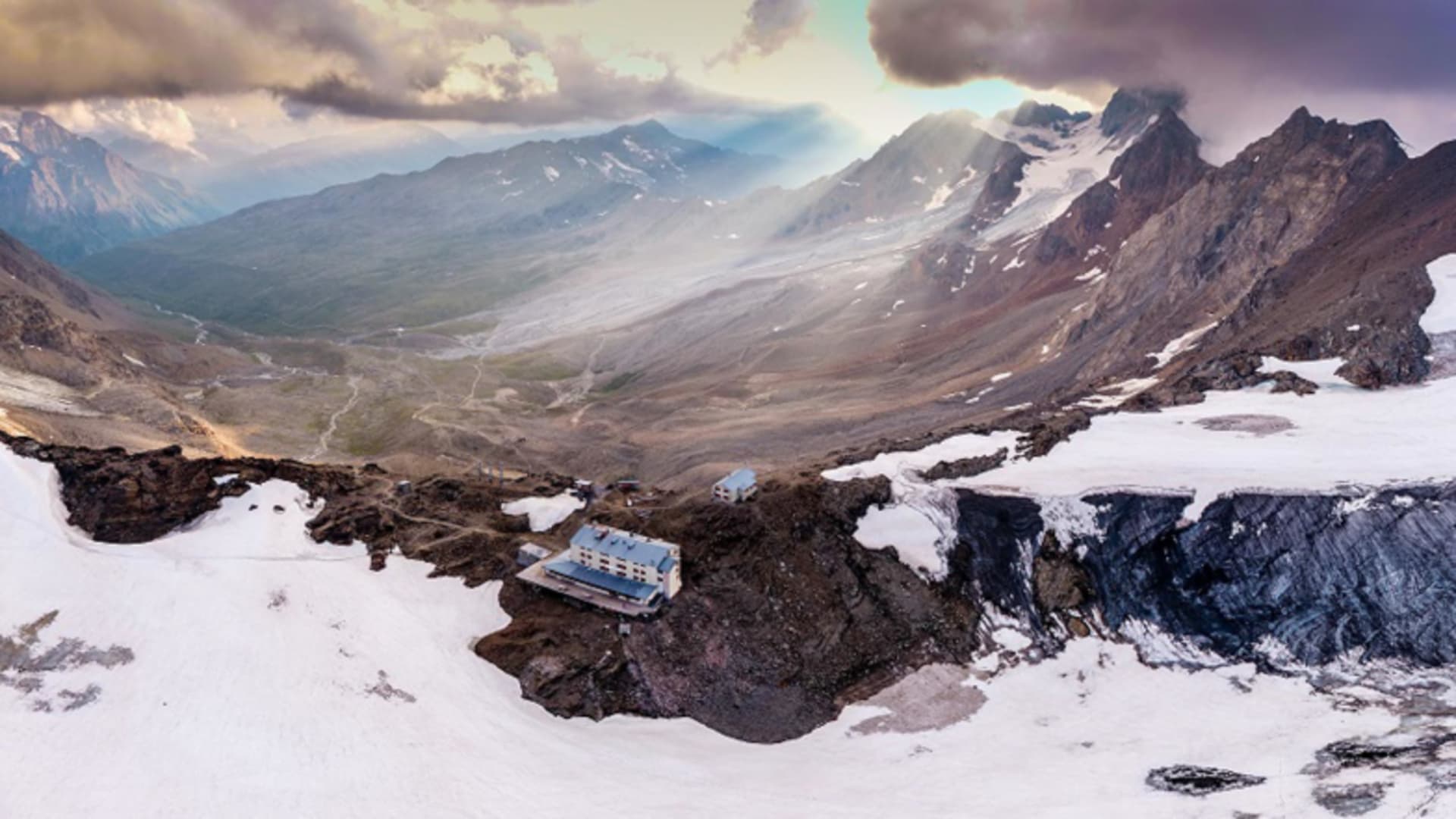 Luftaufnahme zeigt das Rifugio Casati isoliert auf schneefreiem Felsgrat zwischen ausgedehnten Schneeflächen der Hochalp