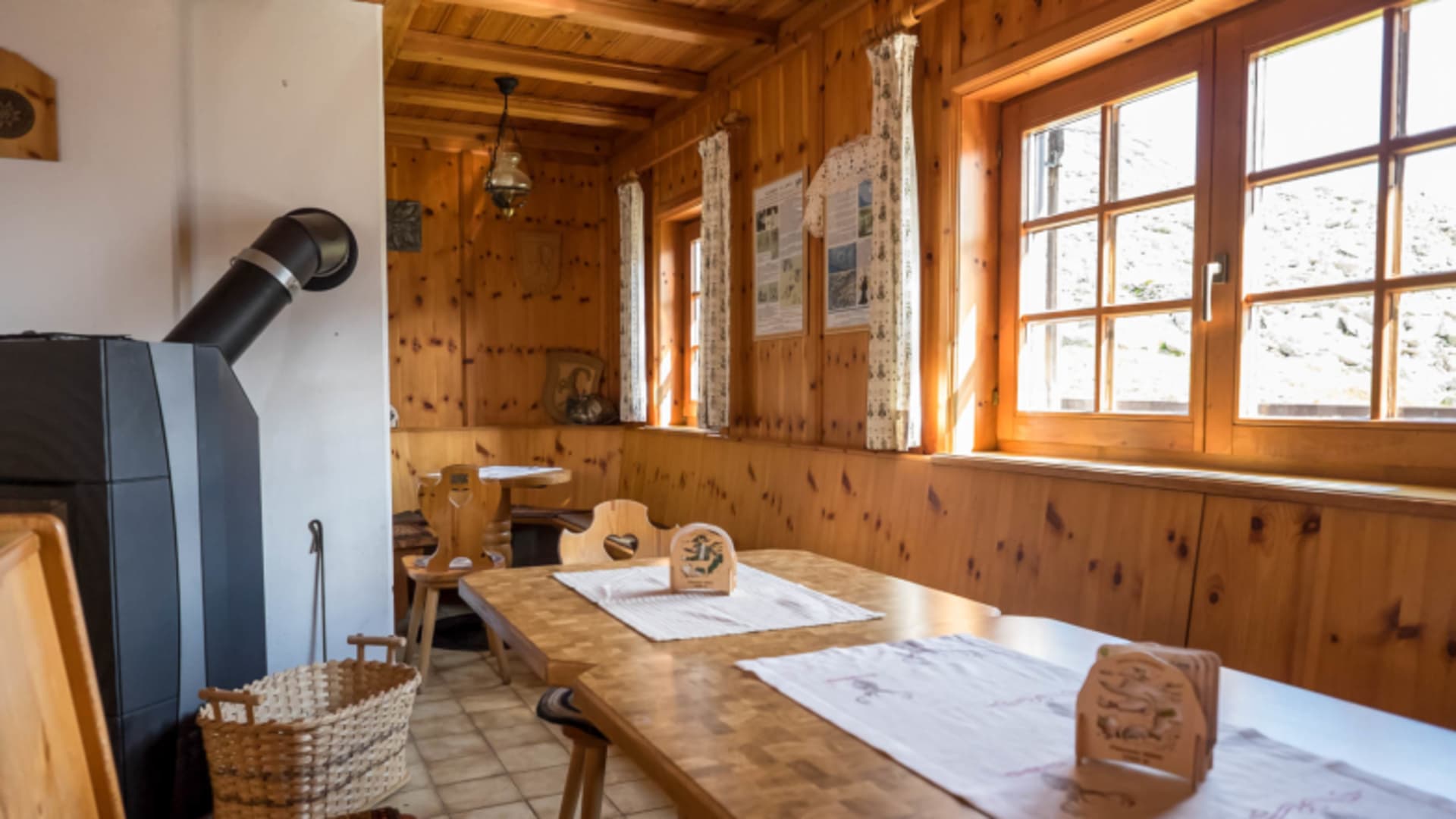 Gastraum der Plauener Hütte mit Holztischen, Kaminofen und Sprossenfenstern.