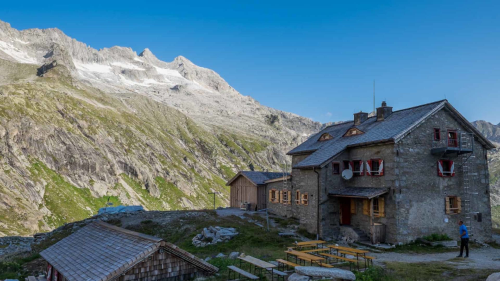 Die Richterhütte mit Außenterrasse und Holzbänken vor der Kulisse schneebedeckter Gipfel.