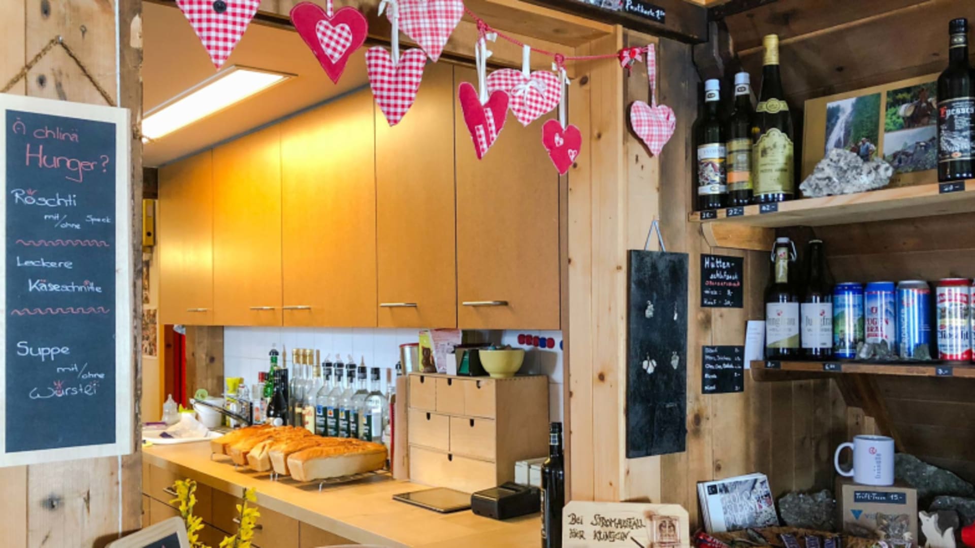 Selbstbedienungsbereich der Trifthütte mit orangefarbenen Küchenschränken, Speisekarte und Getränkeregal mit Holzverklei