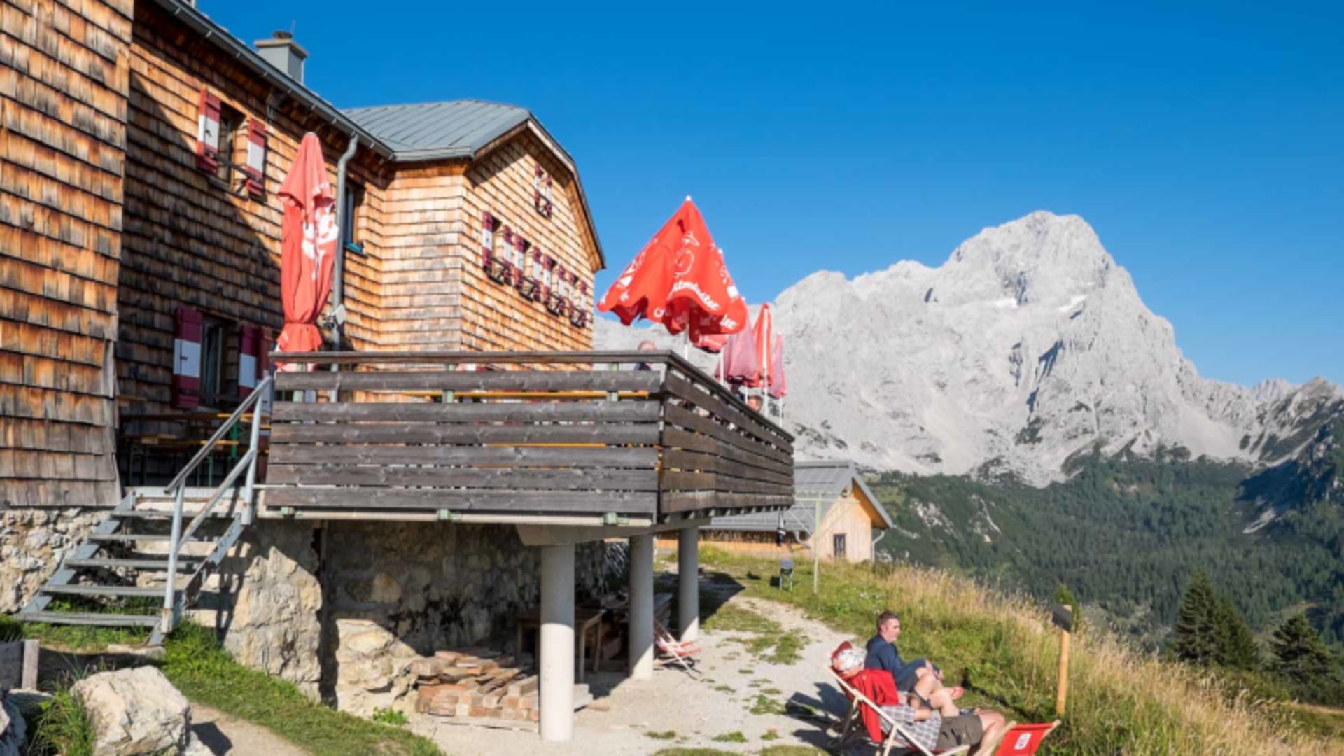Terrasse der Hofpürglhütte mit Gästen in roten Stühlen vor schneebedecktem Gipfel im Hintergrund.