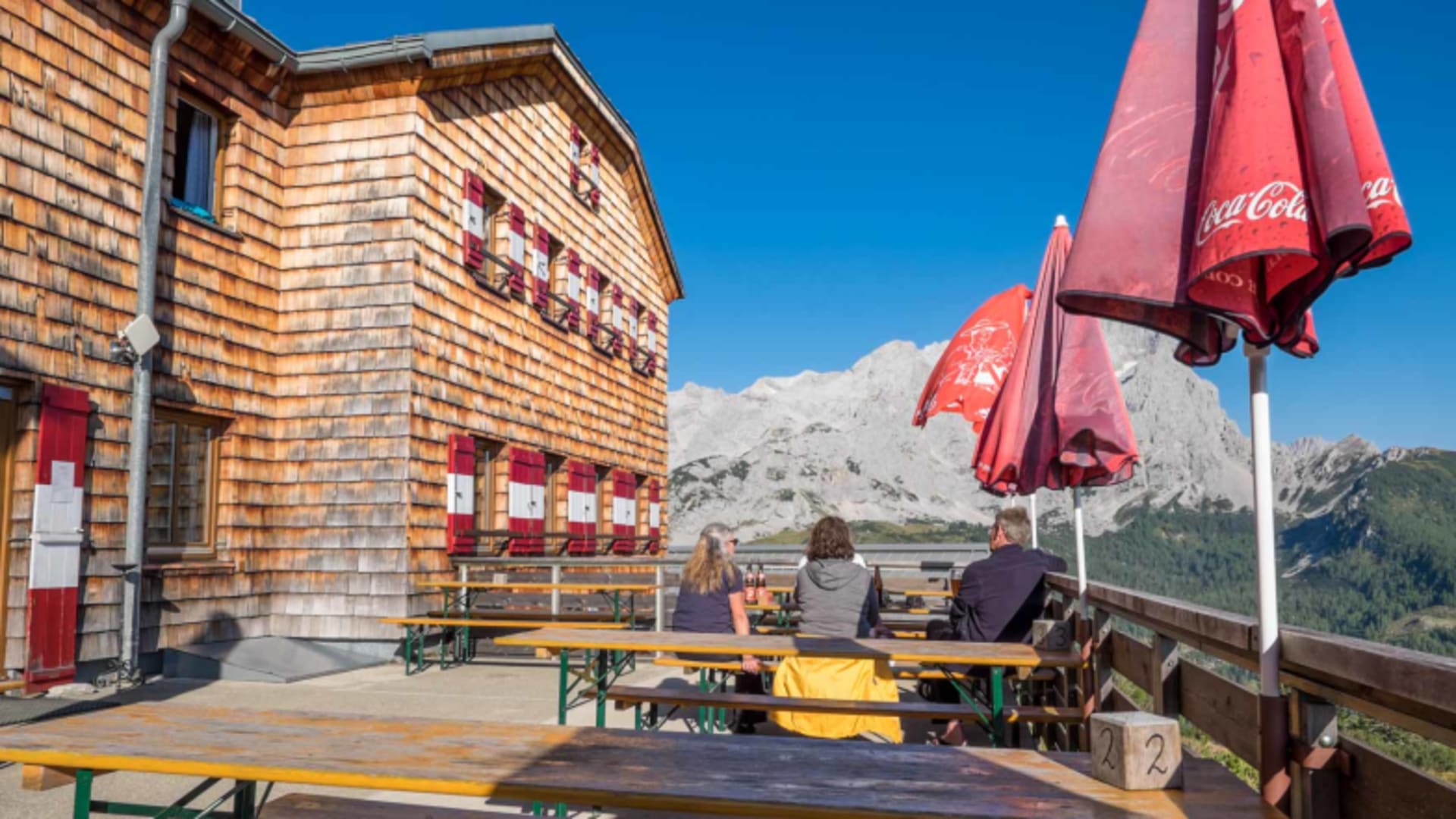 Sonnenterrasse der Hofpürglhütte mit Gästen und Blick auf die Kalksteinfelsen der Bischofsmütze im Hintergrund.