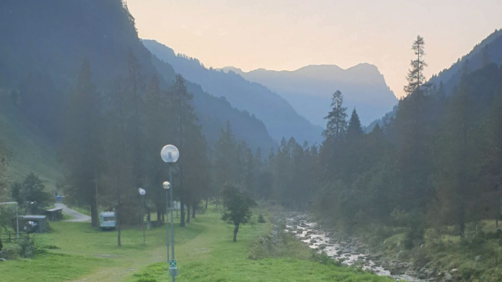 Panorama vom Rifugio Alpenrose über ein grünes Bergtal mit Bach und bewaldeten Hängen bei Sonnenuntergang.