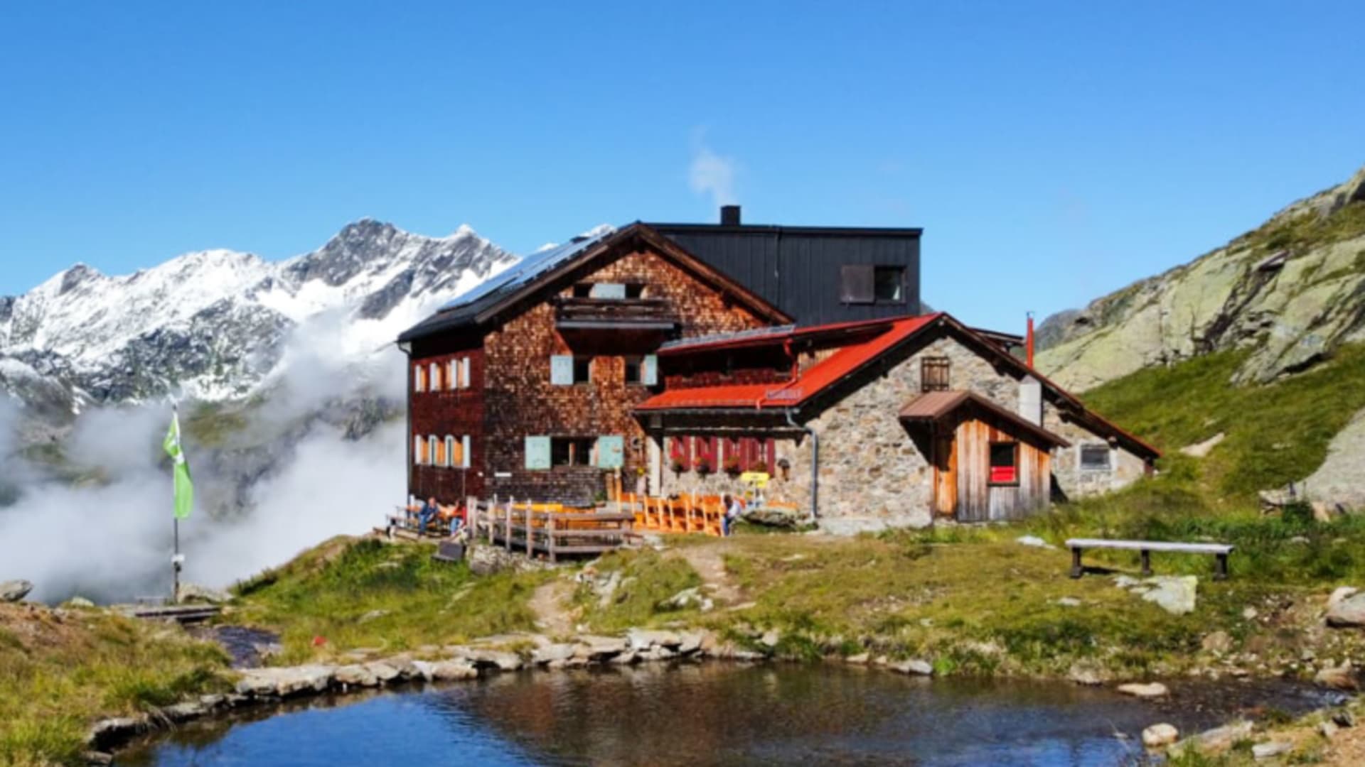 Blick auf die Edmund-Graf-Hütte mit überdachter Terrasse und Gästen am Ufer eines Bergsees bei sonnigem Wetter.