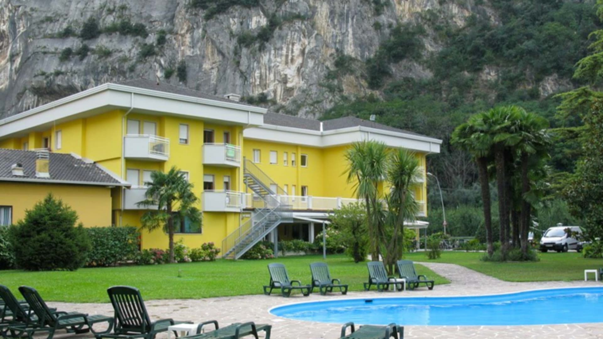 Gelbes Hotelgebäude des Hotel Garden Arco mit rechteckigem Pool und Felswand im Hintergrund.
