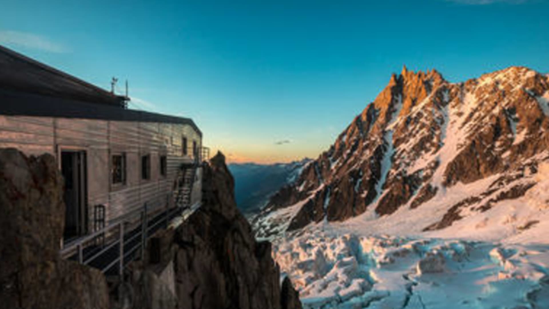 Berghütte Refuge des Grands Mulets auf Felsgrat bei Dämmerung mit verschneiten Gipfeln im Hintergrund.
