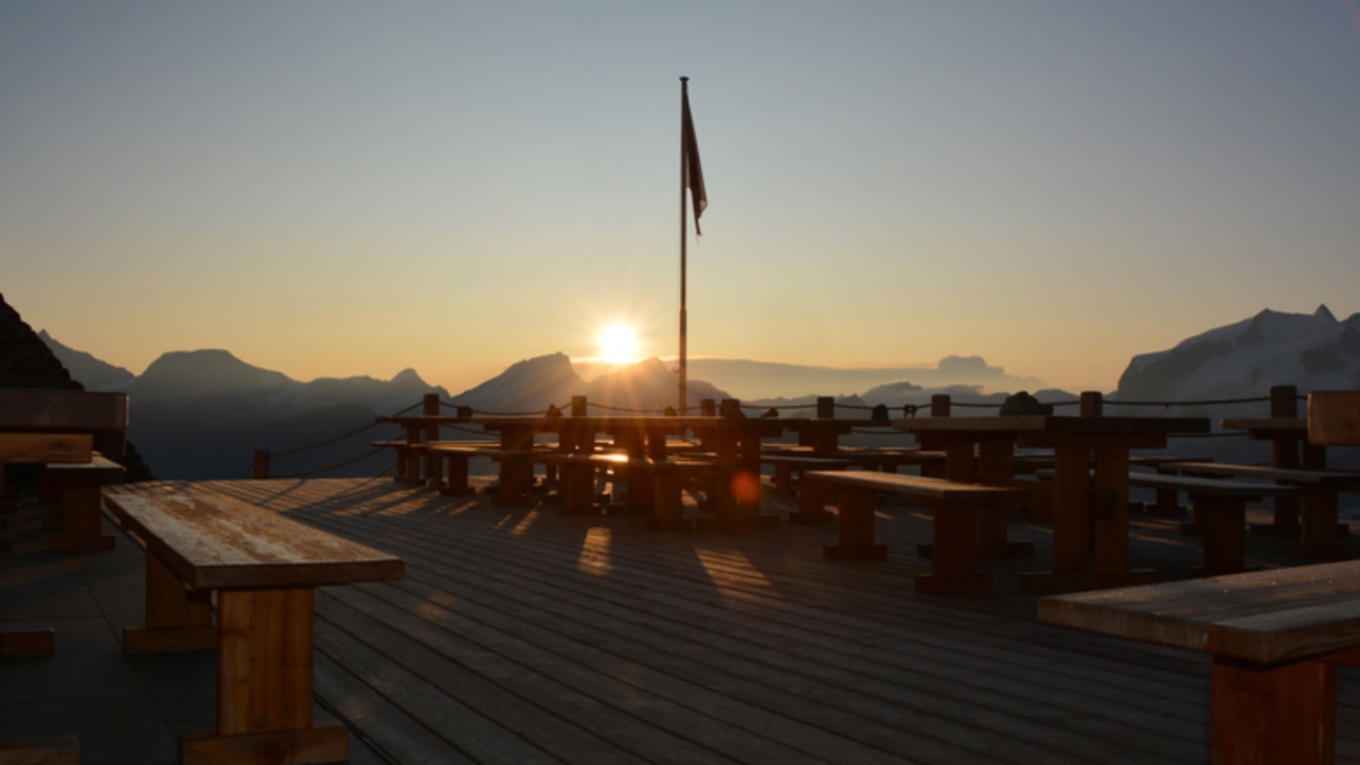 Holzterrasse der Hörnlihütte bei Sonnenuntergang mit Fahnenmast und Bergsilhouette im Gegenlicht.