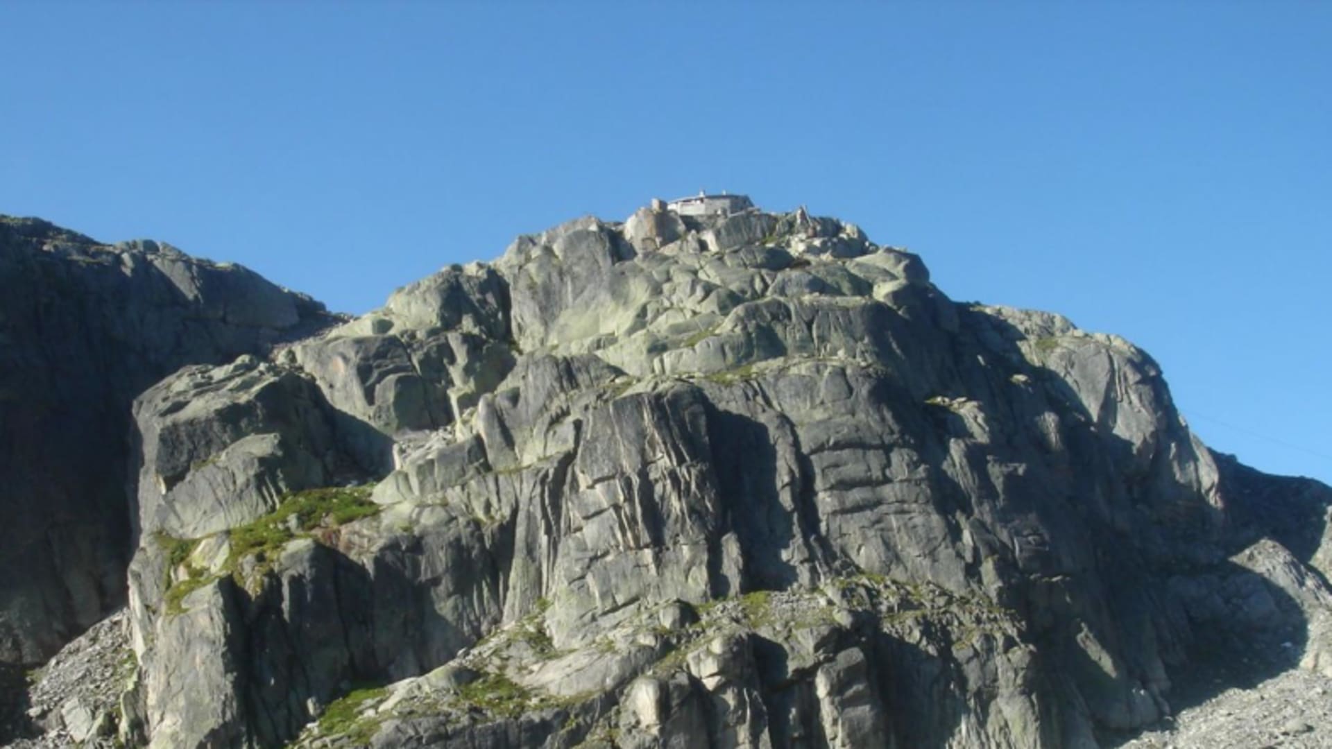 Blick auf die Albert Heim Hütte mit weißem Gebäude und rotem Dach auf einem steilen Kalksteinfelsen.
