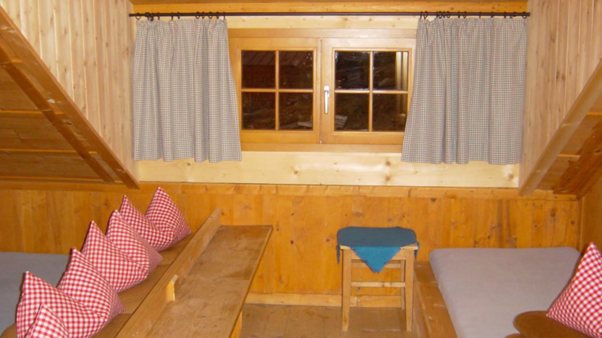 Gemütlicher Aufenthaltsraum der Geraerhütte mit Holz-Eckbank, karierten Kissen und Sprossenfenster.