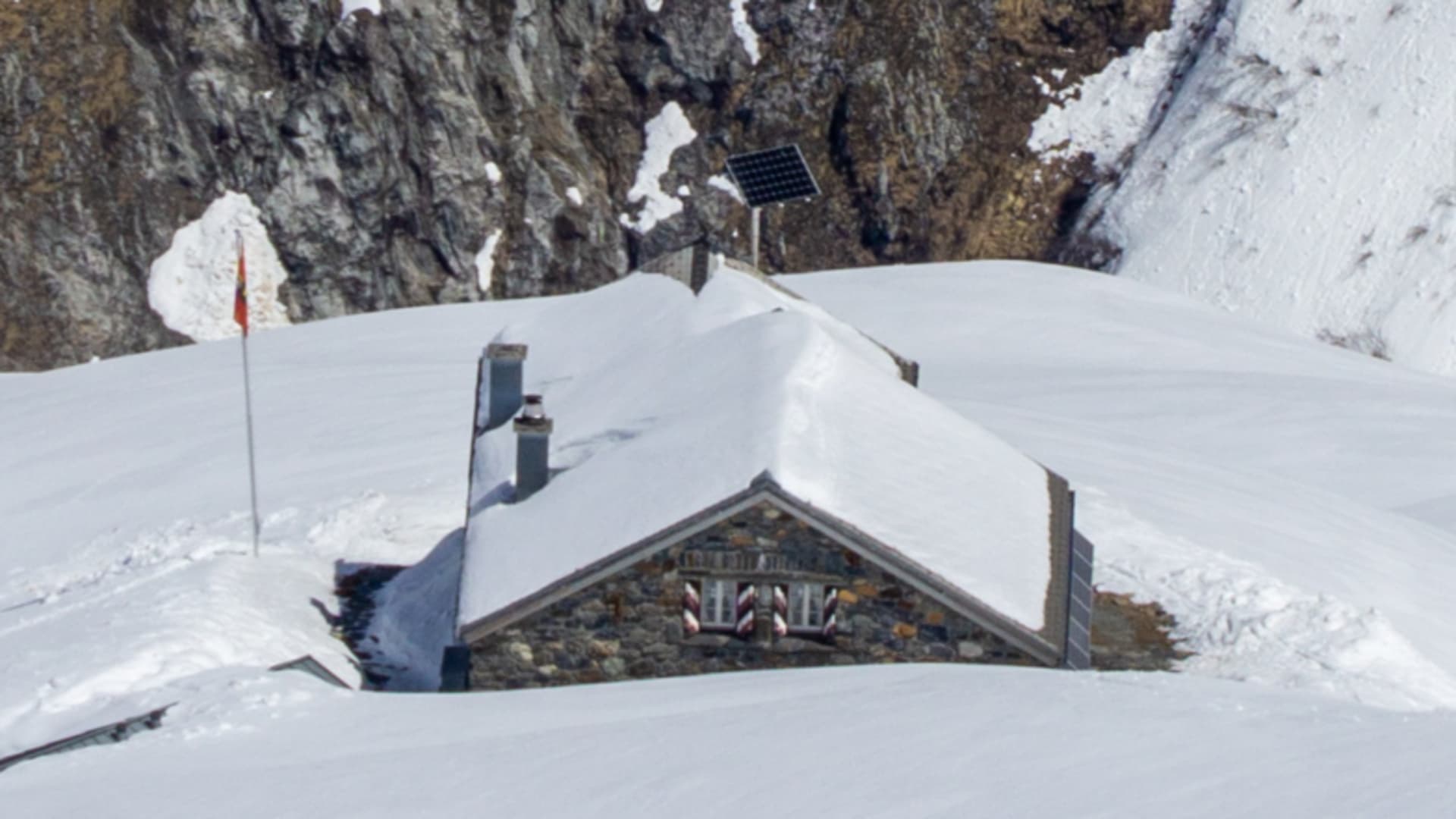 Luftbild der tief verschneiten Fridolinshütte zwischen Felswänden, nur Dach und Schornsteine ragen aus der Schneedecke heraus.