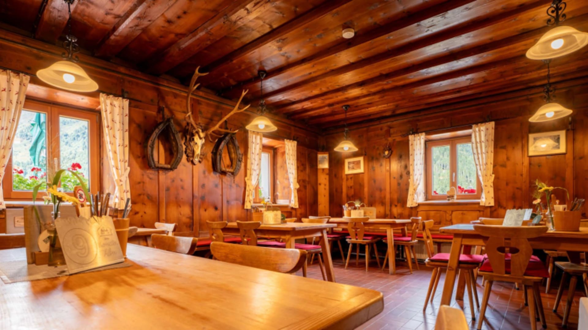 Urige Holzstube der Kemater Alm mit Balkendecke, Hirschgeweih und roten Stuhlpolstern bei warmer Beleuchtung.