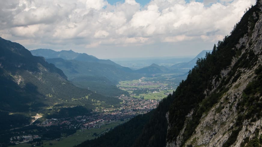 Panoramablick vom Kreuzeckhaus hinab ins Tal nach Garmisch-Partenkirchen mit Siedlung und bewaldeten Berghängen.