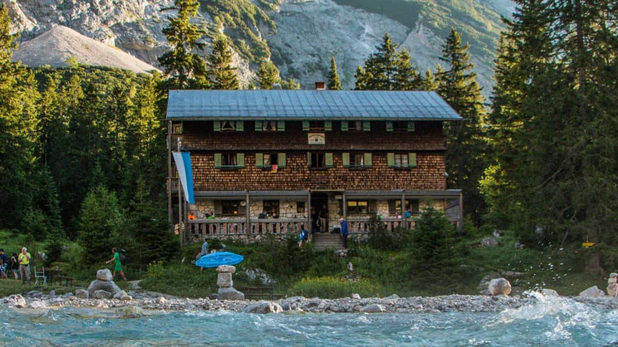 Die Reintalangerhütte im warmen Abendlicht am steinigen Gebirgsbach mit Wanderern auf der Terrasse.