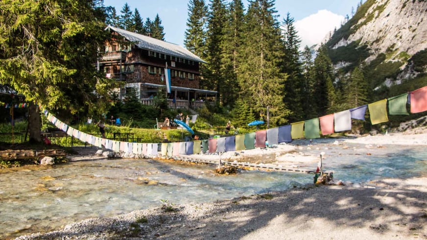 Bergwanderer überqueren die Hängebrücke zur Reintalangerhütte mit bunten Gebetsfahnen über dem türkisen Fluss.