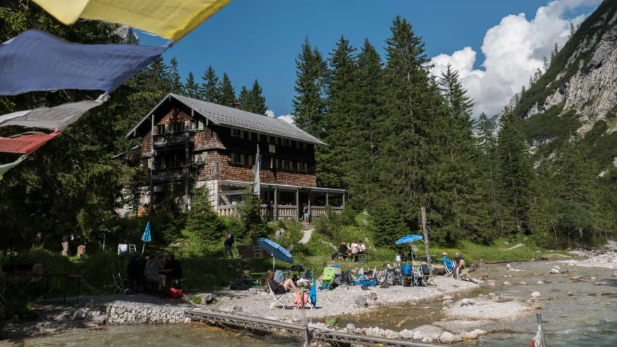 Blick auf die Reintalangerhütte am türkisblauen Gebirgsbach mit bunten Gebetsfahnen zwischen den Bäumen.