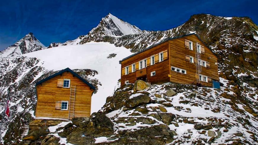 Die Mischabelhütte besteht aus zwei Holzgebäuden auf felsigem Terrain vor schneebedeckten Gipfeln der Walliser Alpen.