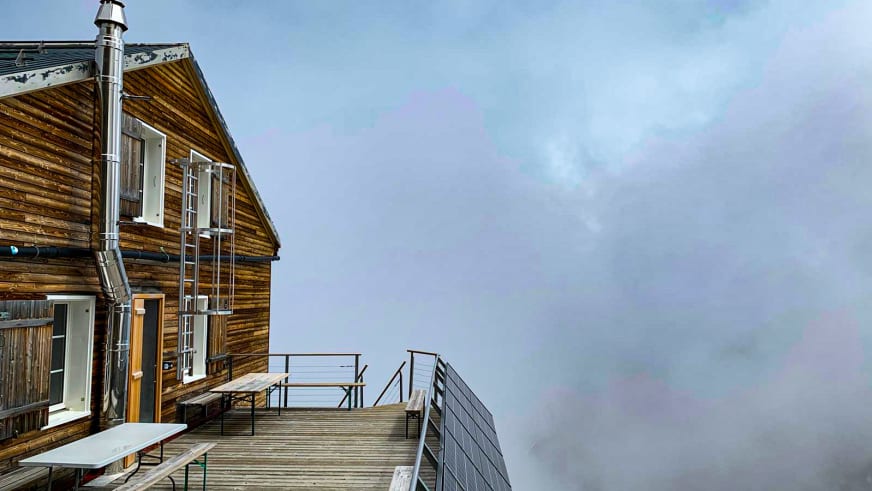 Die Mischabelhütte steht komplett in dichten Wolken gehüllt mit ihrer charakteristischen Holzfassade und Terrasse.