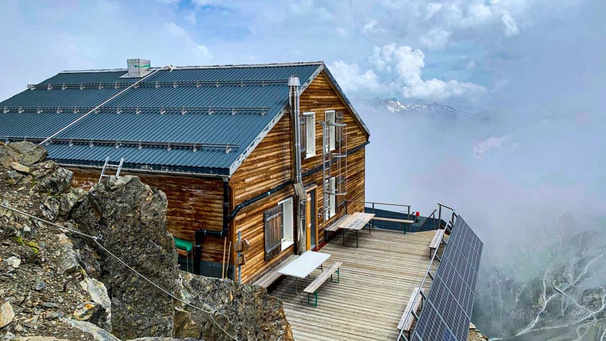 Moderne Mischabelhütte in Holzbauweise mit Solarmodulen auf der Terrasse vor schneebedeckten Berggipfeln der Walliser Al