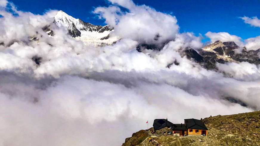 Panoramablick auf die Domhütte über dichter Wolkendecke mit schneebedeckten Gipfeln im Hintergrund.