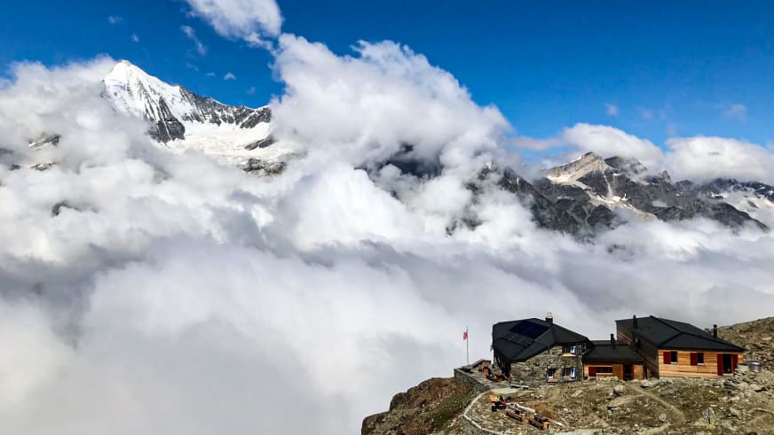 Die Domhütte steht auf einem Felsplateau über dichter Wolkendecke mit Blick auf schneebedeckte Gipfel.