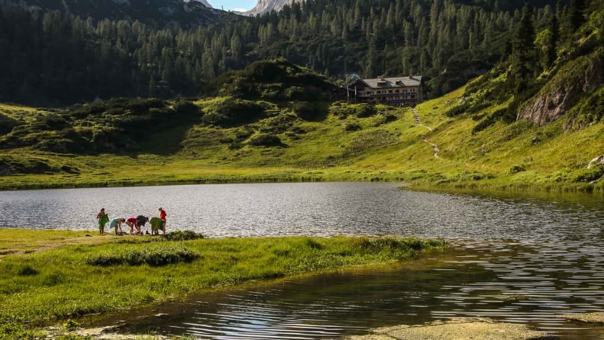 Fünf Wanderer pausieren am Ufer des Funtensees mit Blick auf das Kärlingerhaus am bewaldeten Berghang.