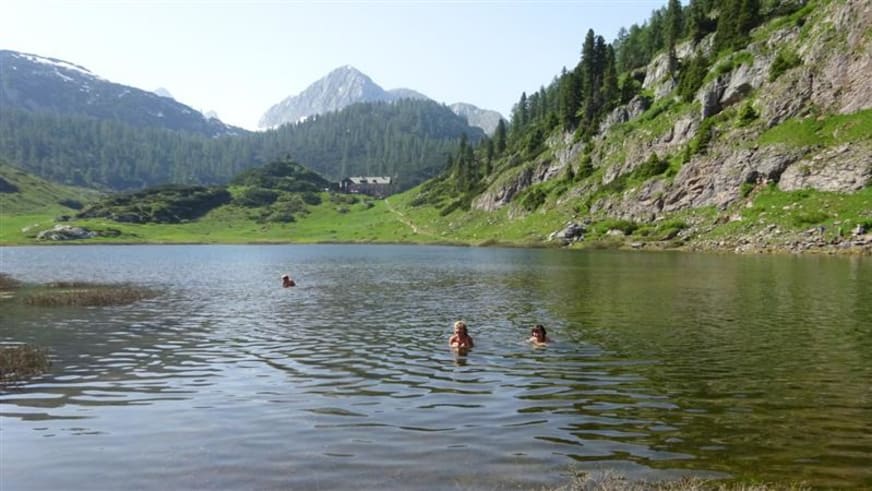 Drei Personen baden im klaren Bergsee nahe dem Kärlingerhaus mit schneebedeckten Gipfeln und bewaldeten Hängen im Hintergrund.