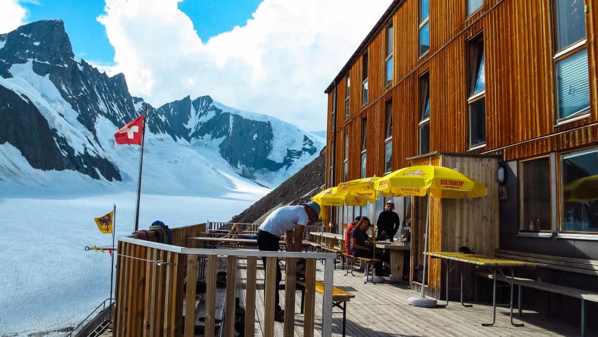Gäste entspannen unter gelben Sonnenschirmen auf der Holzterrasse der Finsteraarhornhütte vor Gletscherkulisse.
