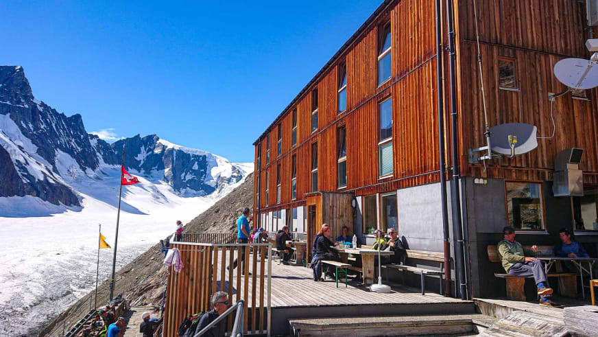 Etwa 15 Personen pausieren an Holztischen auf der Terrasse der Finsteraarhornhütte vor Gletscherkulisse.