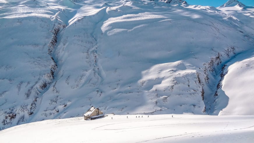 Winteraufnahme der Vernagthütte mit Skifahrern auf verschneiter Fläche vor Bergkulisse der Ötztaler Alpen.