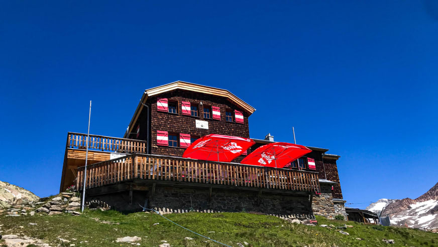 Außenansicht der Vernagthütte mit großer Holzterrasse und roten Sonnenschirmen vor Bergpanorama.