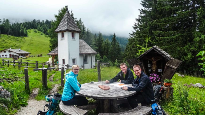 Drei Bergwanderer pausieren am Holztisch vor der weißen Kapelle der Kührointhütte.