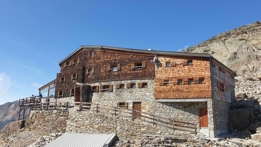 Außenansicht der Similaunhütte auf 3.019 m Höhe mit Holzterrasse und Bergsteigern in hochalpiner Umgebung.