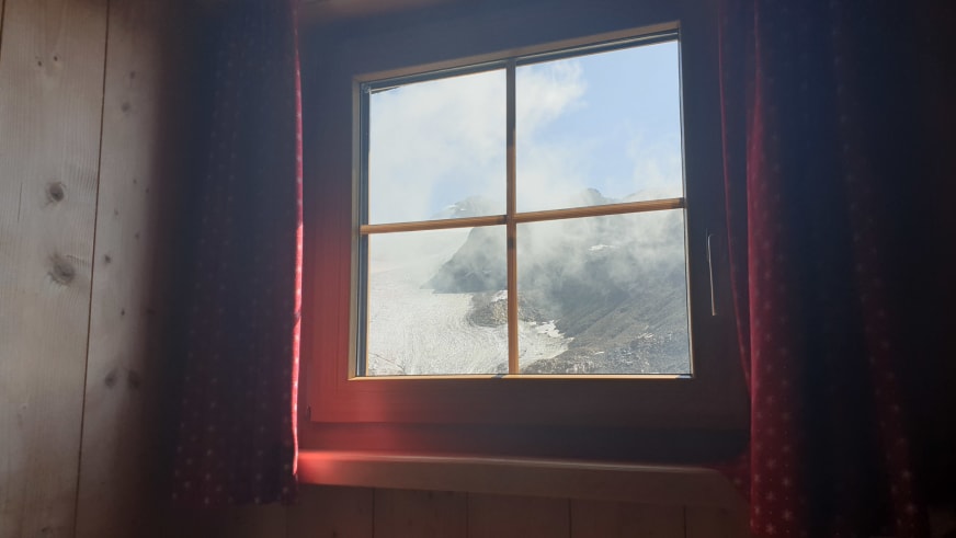 Blick aus einem Zimmer der Similaunhütte durch vierteiliges Holzfenster auf die verschneite Gletscherlandschaft.